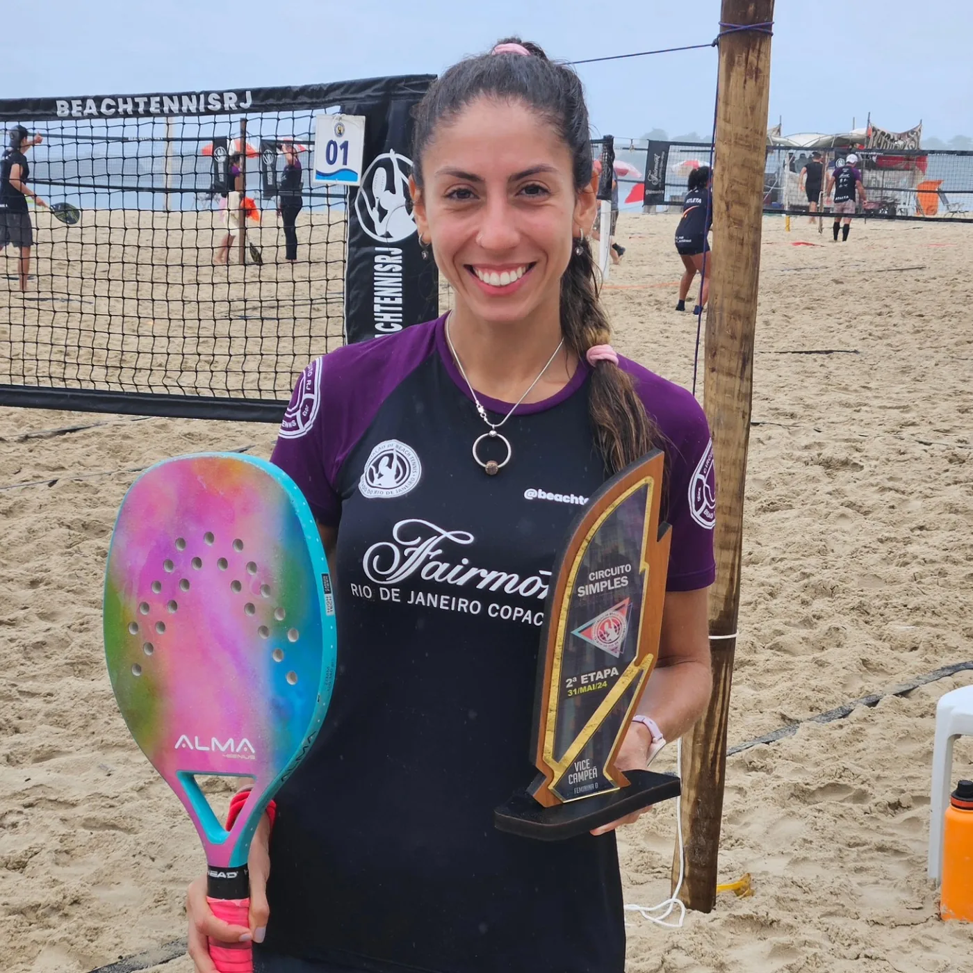 Carol segurando troféu e raquete em quadra de beach tennis