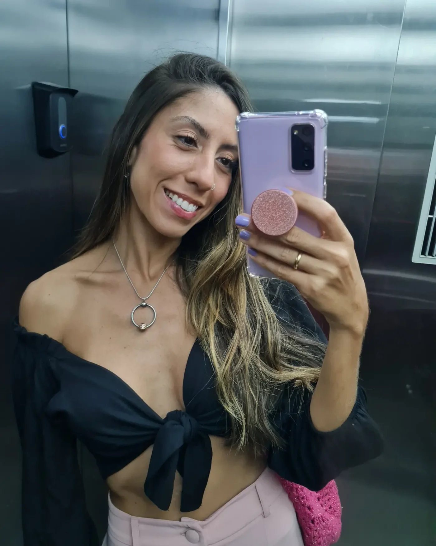 Carol tirando selfie no espelho do elevador, de blusa preta