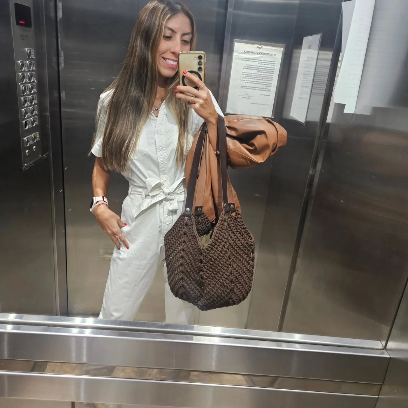 Carol de roupa branca tirando selfie no espelho do elevador com uma bolsa de crochê