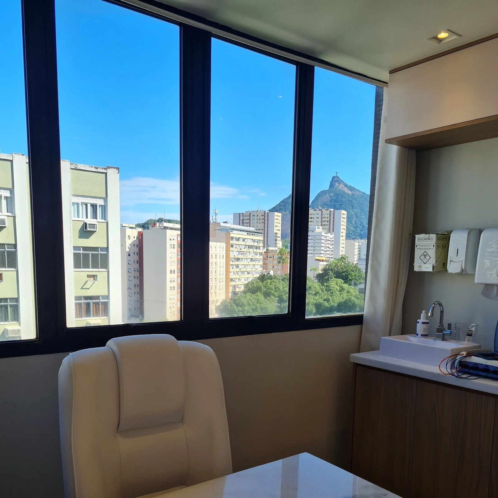 Sala da clínica com poltrona creme junto à janela com vista direta para o Cristo Redentor e os prédios do Flamengo